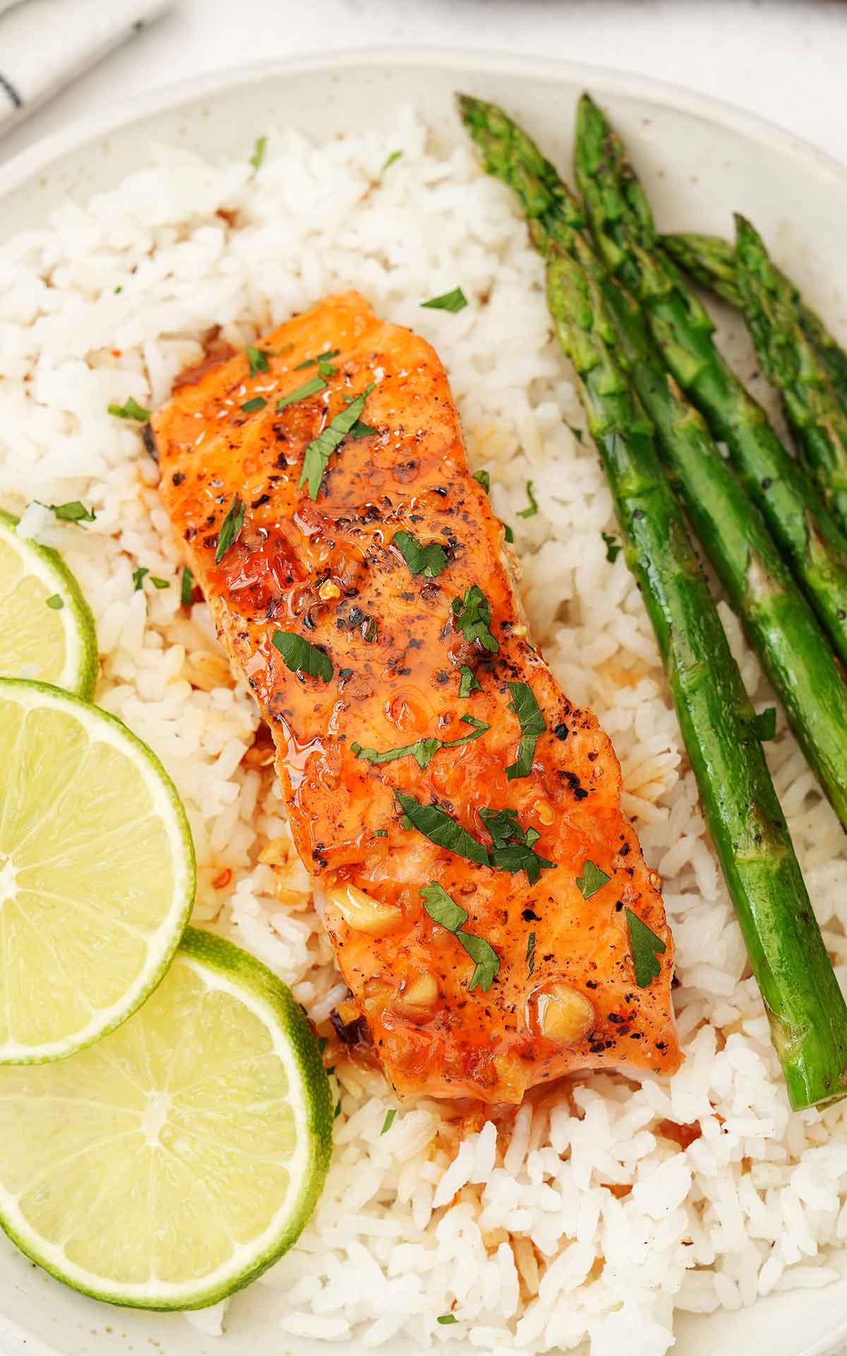 A white plate containing rice and sweet chilli glazed salmon with asparagus and lime wedges on the side.