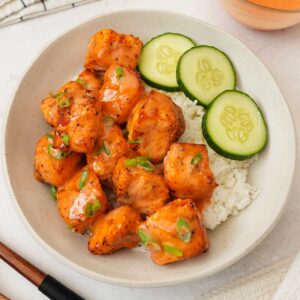 A white rice bowl containing bang bang salmon bites with cucumber slices on the side.
