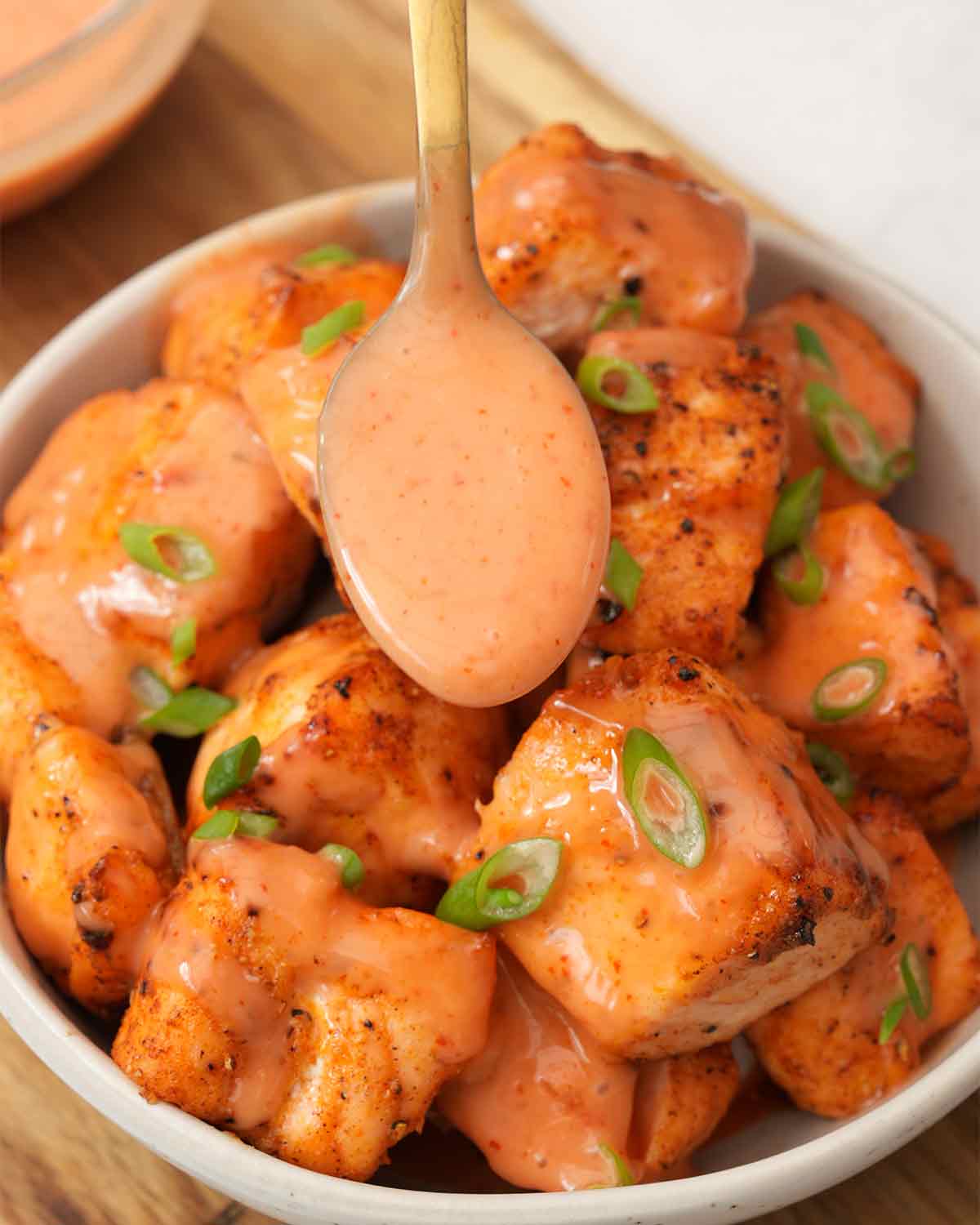 An image showing a bowl of fried salmon pieces with a teaspoon on top drizzling bang bang sauce.