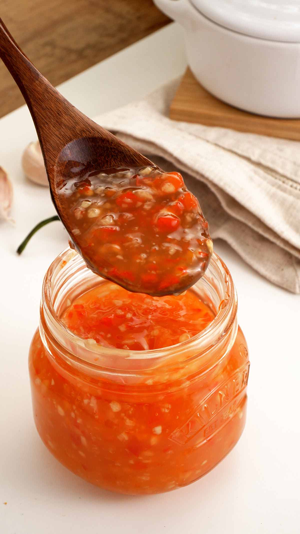 A wooden spoon and a glass jar containing thick glossy sweet chilli sauce. 