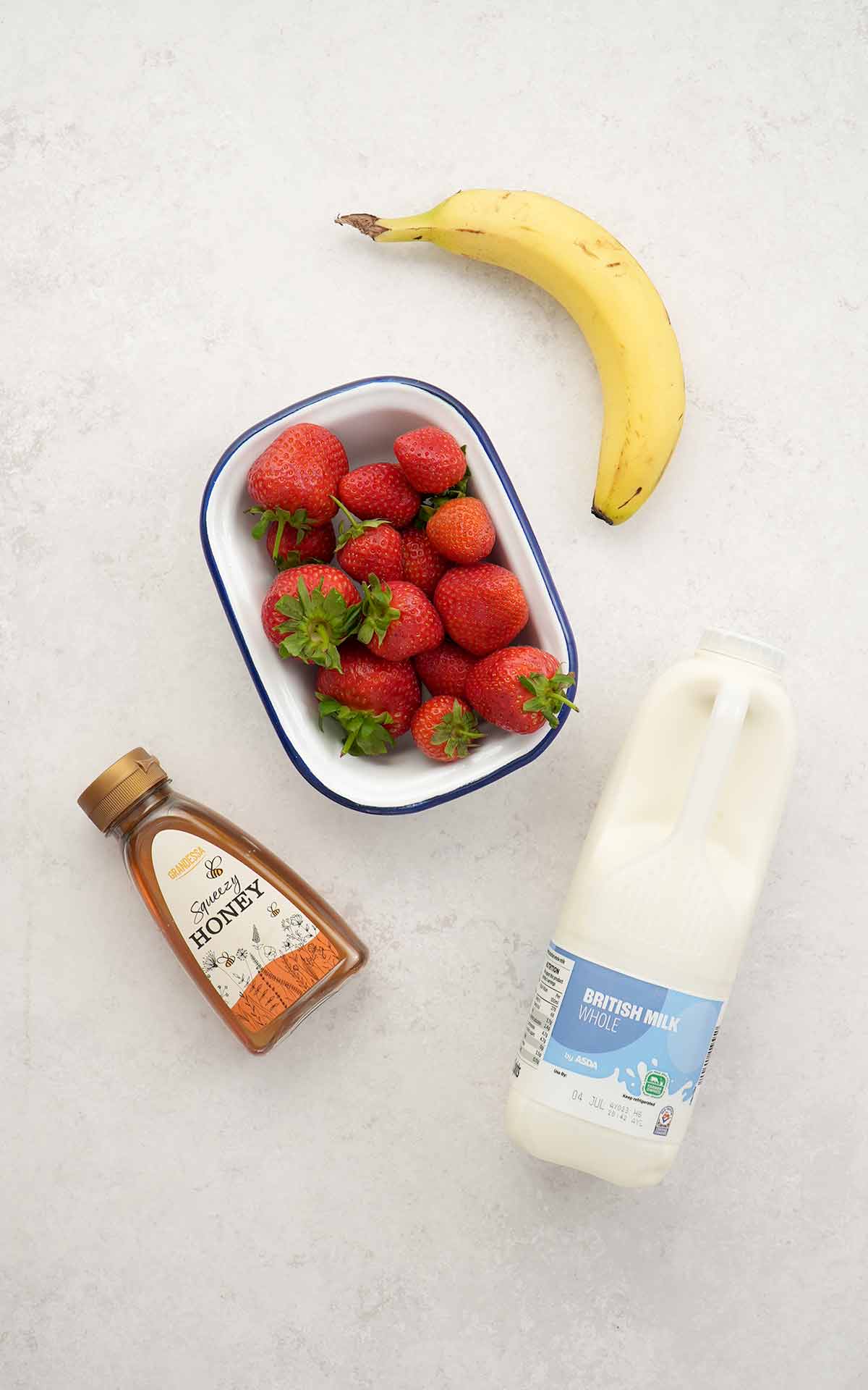 Ingredients of making strawberry and banana milkshake: fresh strawberries, one banana, honey bottle, and milk bottle displayed on the white table. 