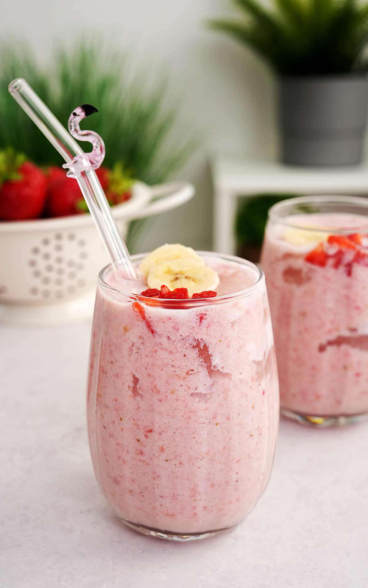 An image of clear glass containing banana and strawberry milkshake, decorated with glass straw and strawberries and banana chunks. 
