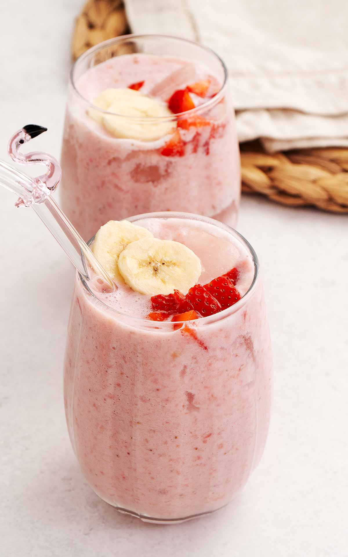 Light pink colour strawberry and banana milkshake in a clear glass. Garnished with slices of banana and strawberries.