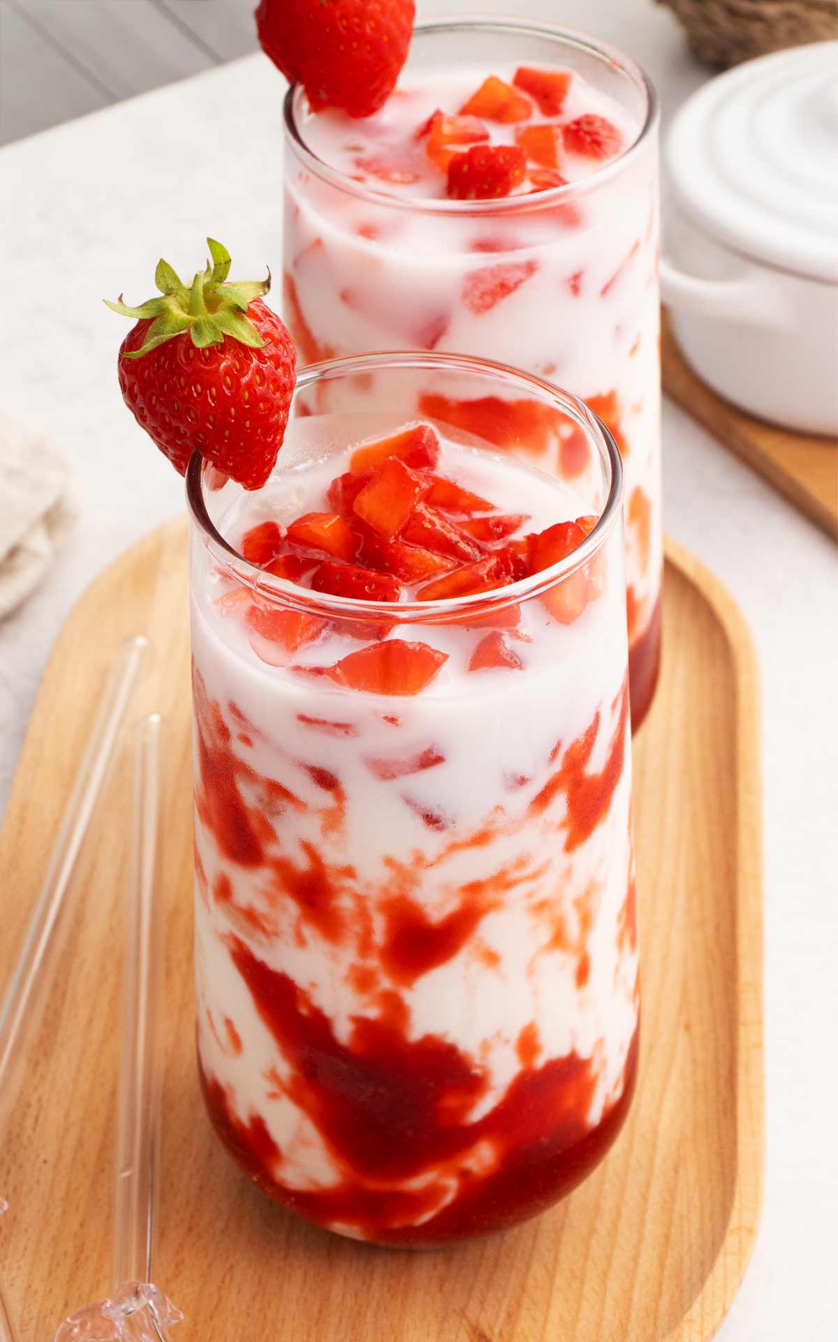 A clear glass containing red strawberry syrup at the bottom layer, white milk in the middle, and small strawberry chunks on top. 