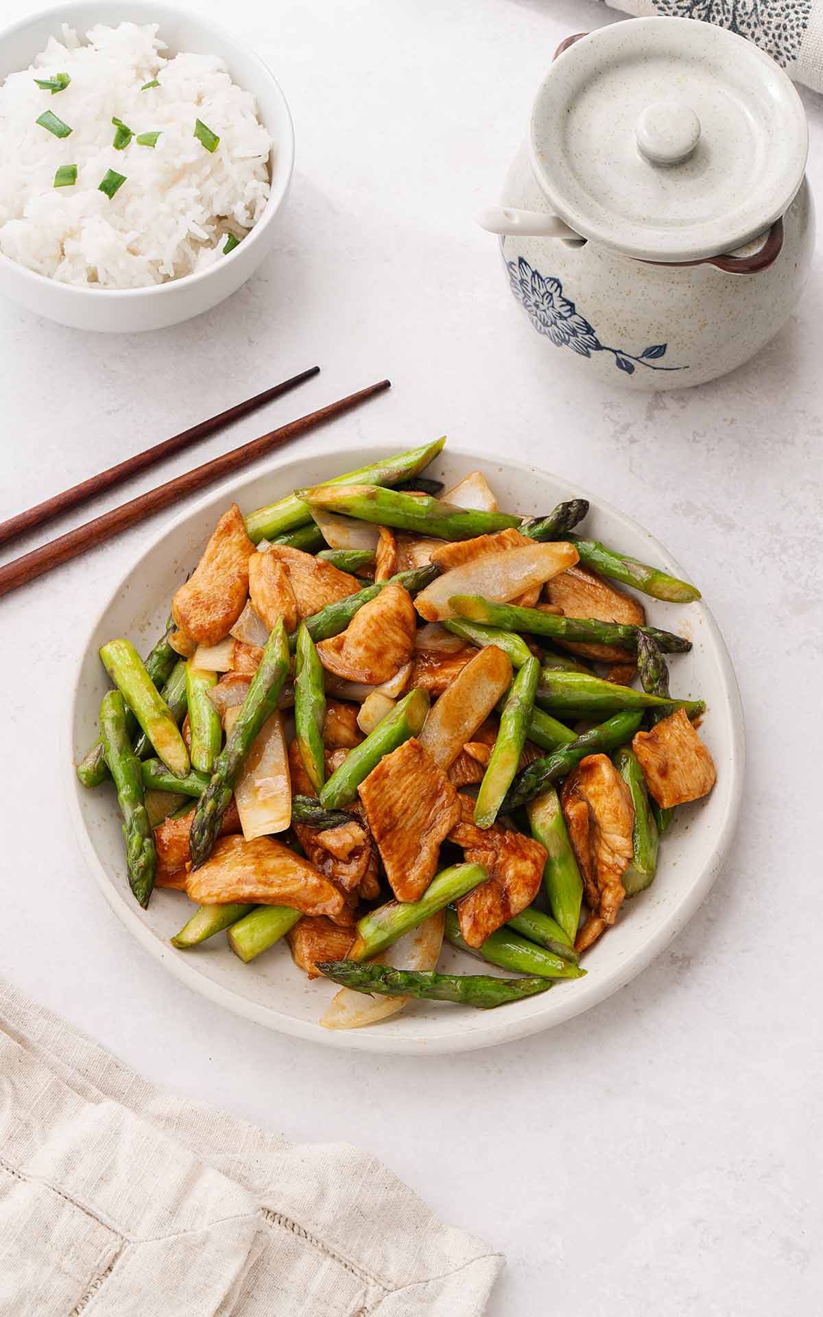 A photo of a white plate containing asparagus and chicken stir fry with rice bowl on the side. 