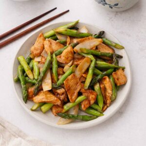 A white plate containing stir fried asparagus chicken dish with wooden chopsticks on the side.