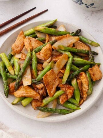 A white plate containing stir fried asparagus chicken dish with wooden chopsticks on the side.