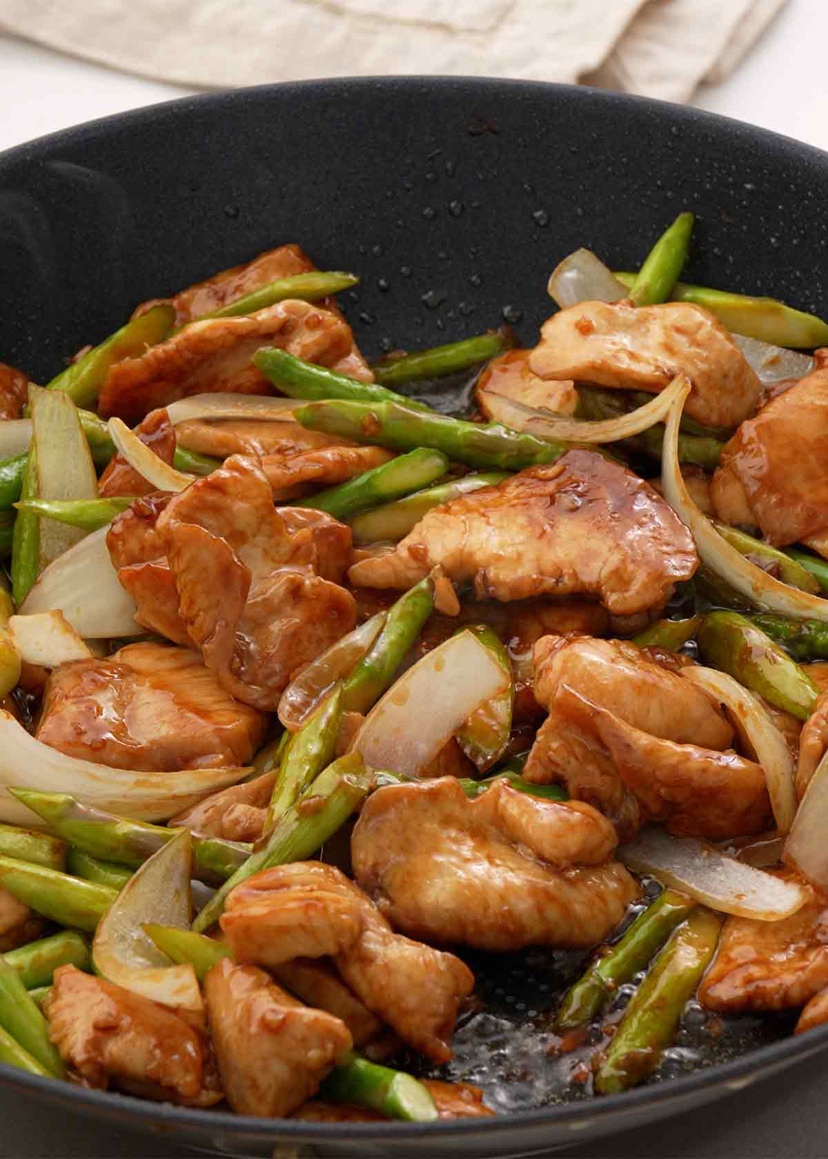 An image showing how to stir fry chicken and asparagus in a large black wok. 