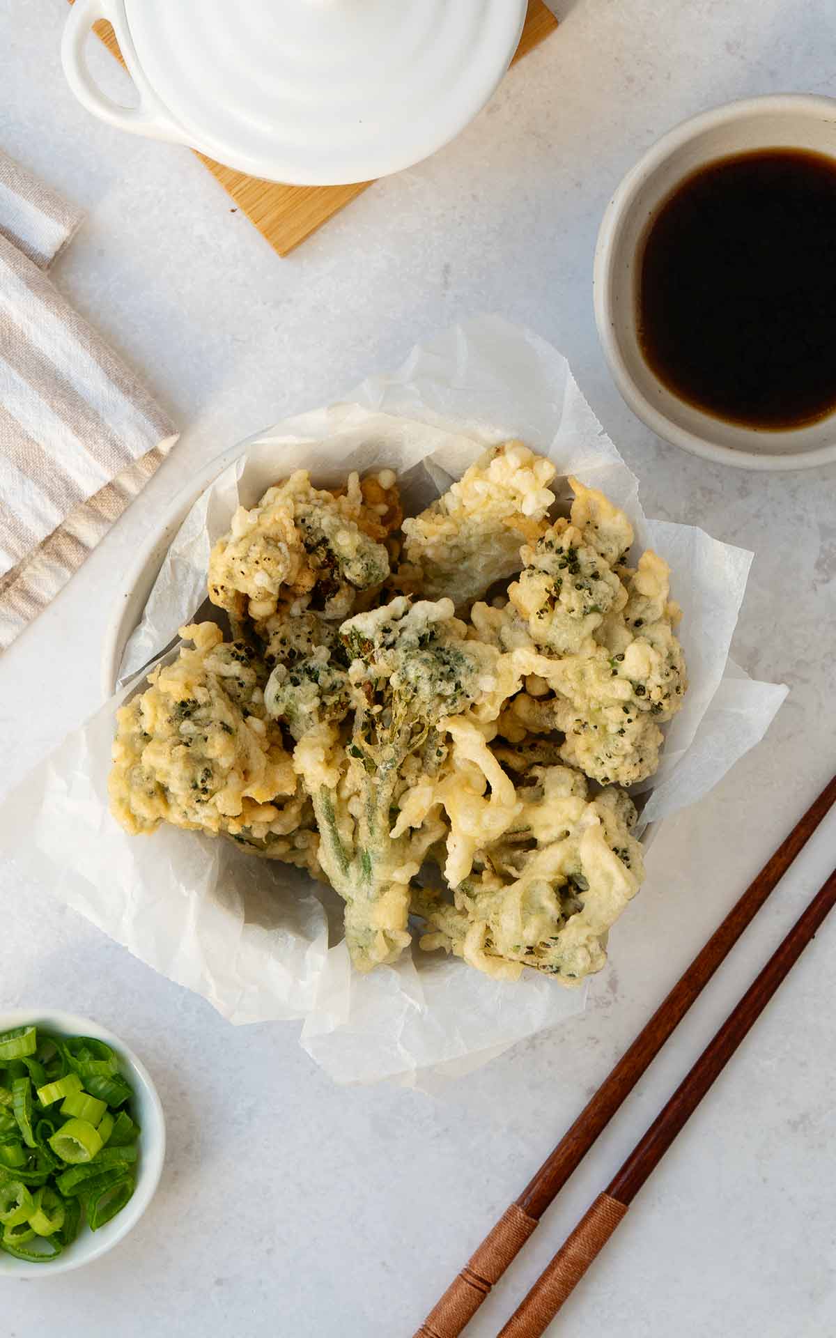 A white bowl containing crispy fried tempura battered coated broccoli bites. Small soy sauce dipping sauce bowl, chopsticks and chopped spring onions on the side.