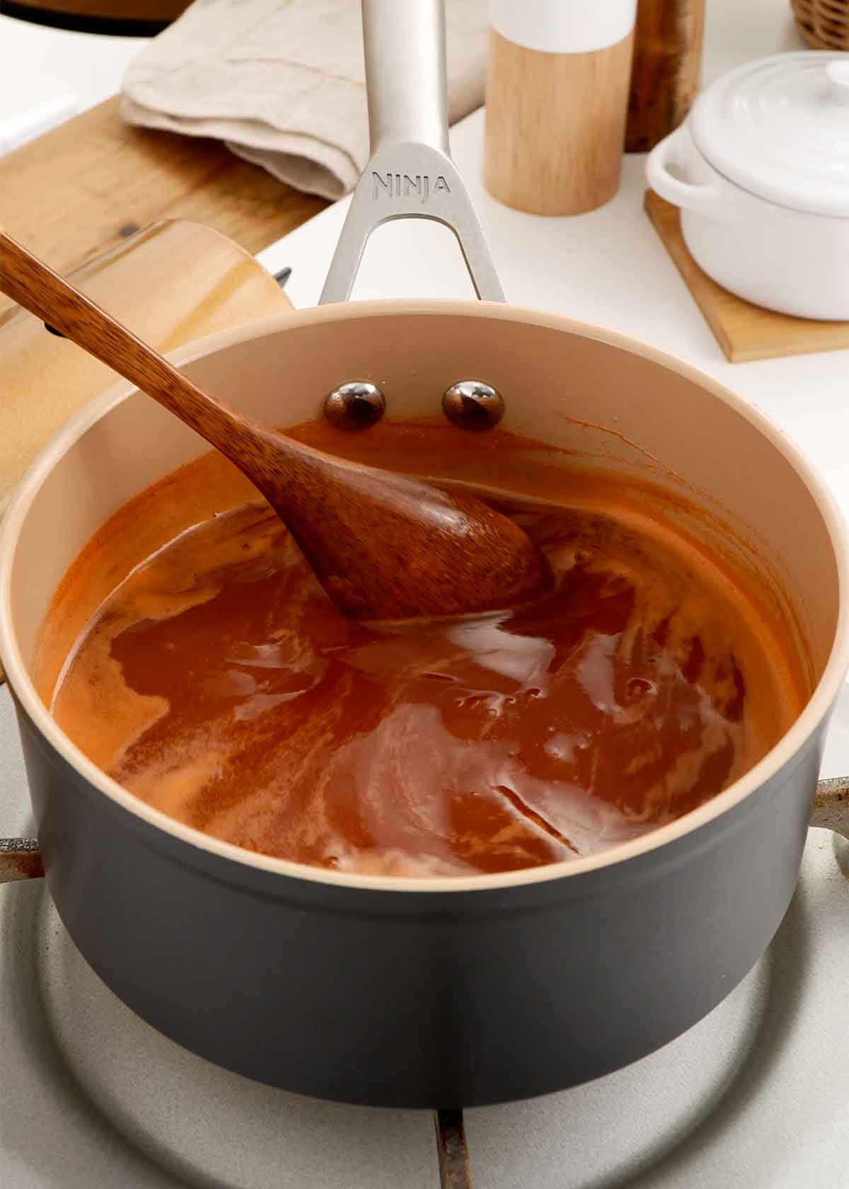 A picture of a sauce pan containing red colour sweet and sour sauce and a wooden spoon is stirring the sauce.