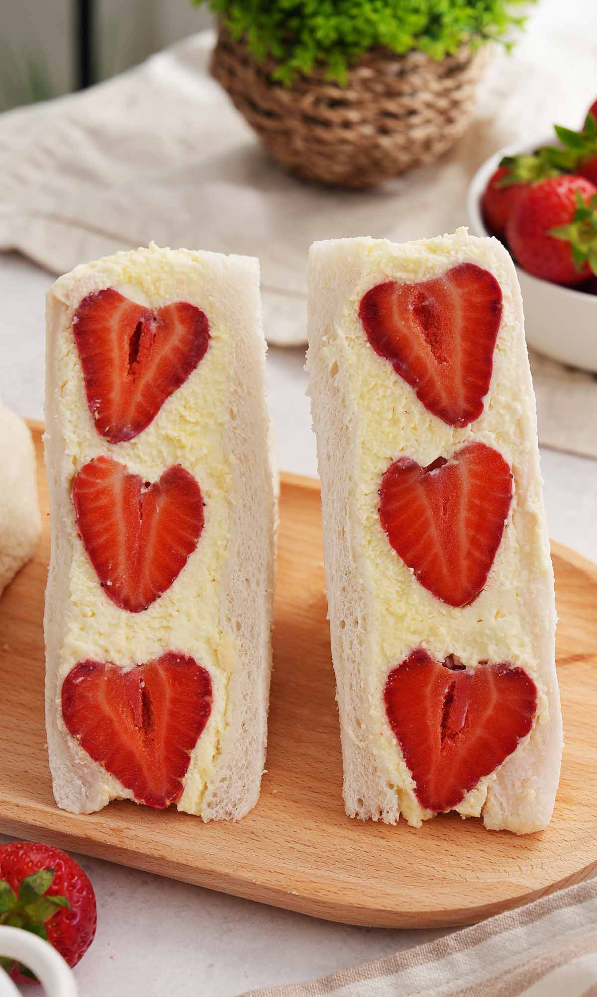 An image showing two halves Japanese strawberry cream sandwiches with thick cream and sliced strawberries on wooden plate.