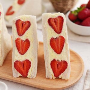 A picture of two half Japanese strawberry sandwiches with large red strawberries and white whipped cream fillings, displayed on a wooden serving plate.