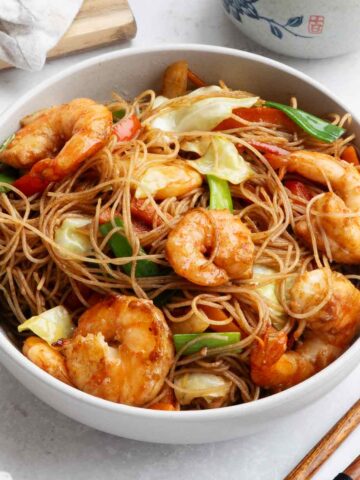 A white bowl containing mei fun noodles with shrimp, cabbage, carrot, and colourful vegetables.