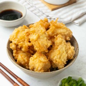 Fried golden tempura fish bites displayed in a brown bowl. A pair of wooden chopsticks, green onions, and soy dipping sauce on the side.