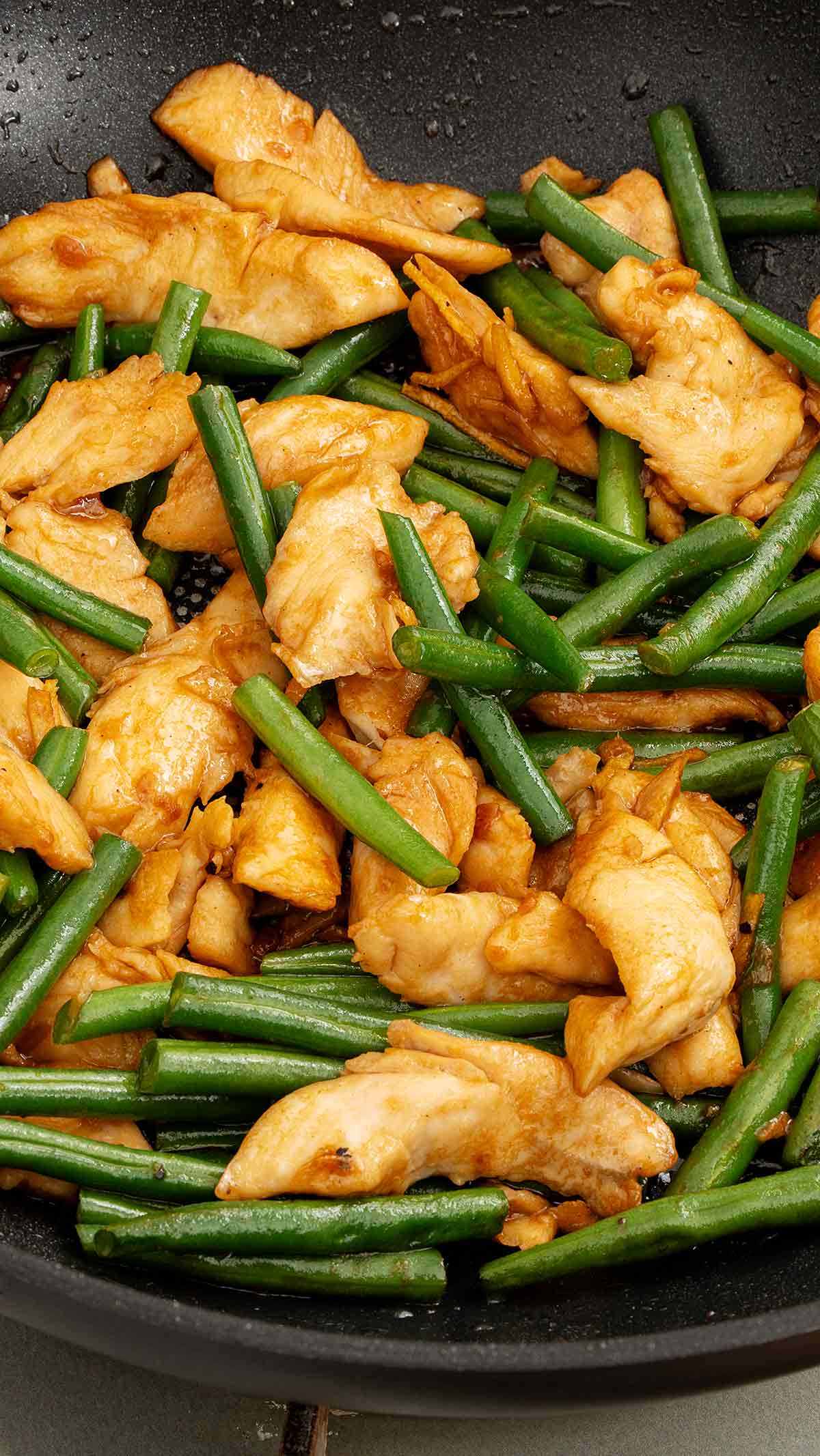 A large black wok containing chicken slices with bright green beans, garlic and ginger.