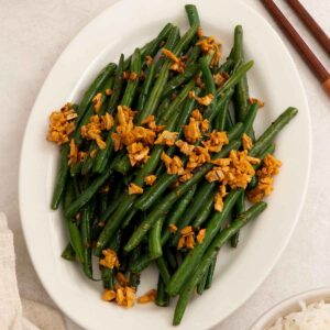 A white plate containing stir fried green beans with crispy golden garlic.