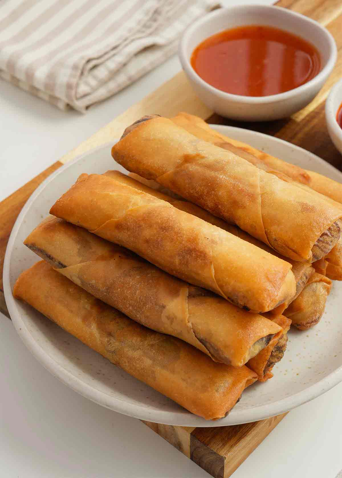 A stack of fried golden spring rolls displayed on the white plate. Red sweet and sour dipping sauce bowl on the side.