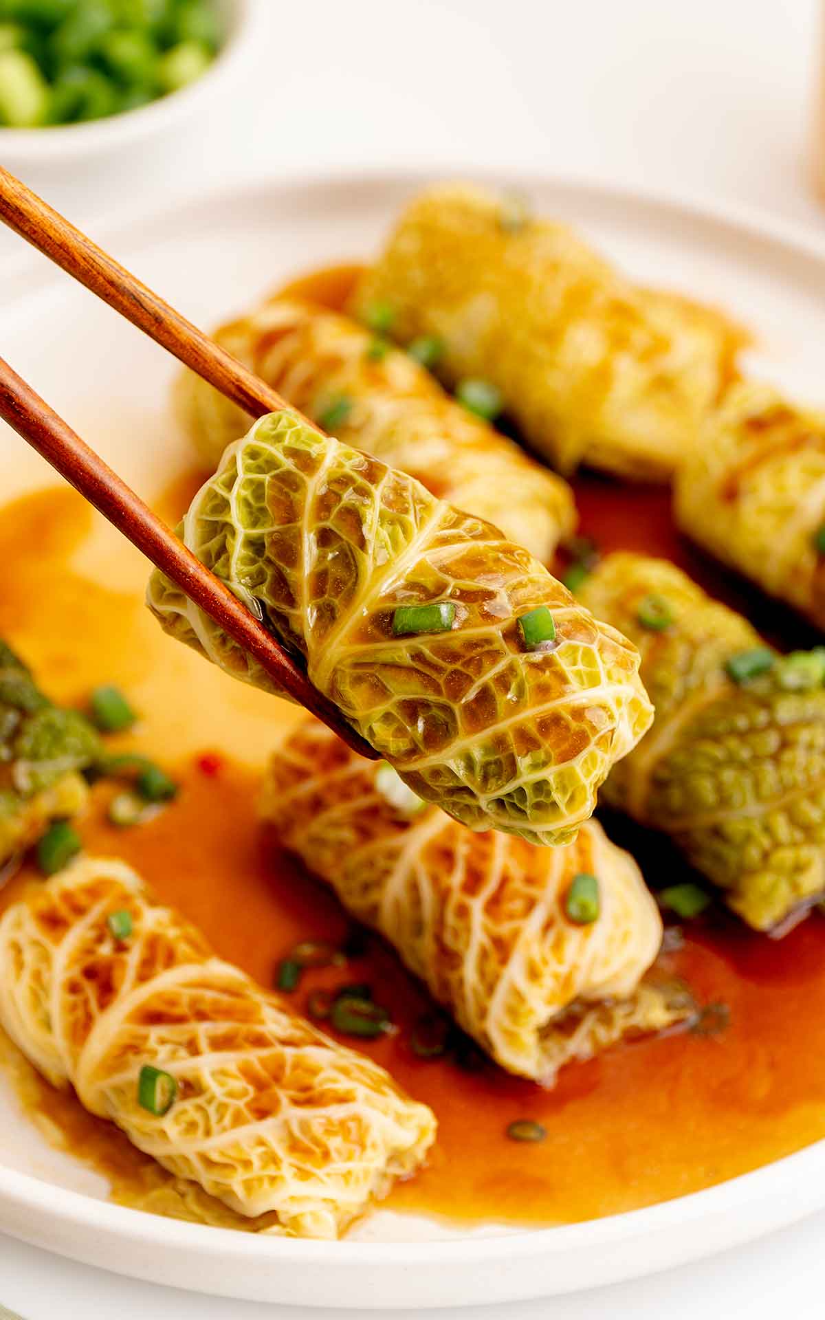 A pair of wooden chopsticks holding steamed cabbage rolls with glossy Chinese brown sauce. Garnish with green onions slices.