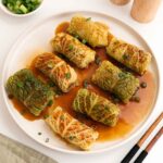 A white plate containing steamed Chinese cabbage rolls with glossy brown sauce and garnish with green onions.