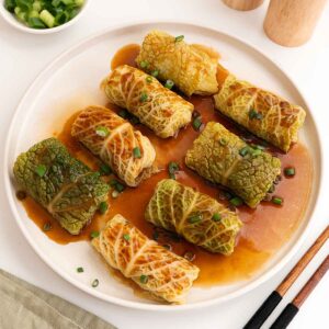 A white plate containing steamed Chinese cabbage rolls with glossy brown sauce and garnish with green onions.
