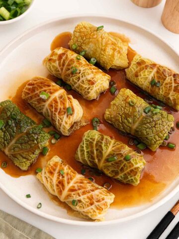 A white plate containing steamed Chinese cabbage rolls with glossy brown sauce and garnish with green onions.