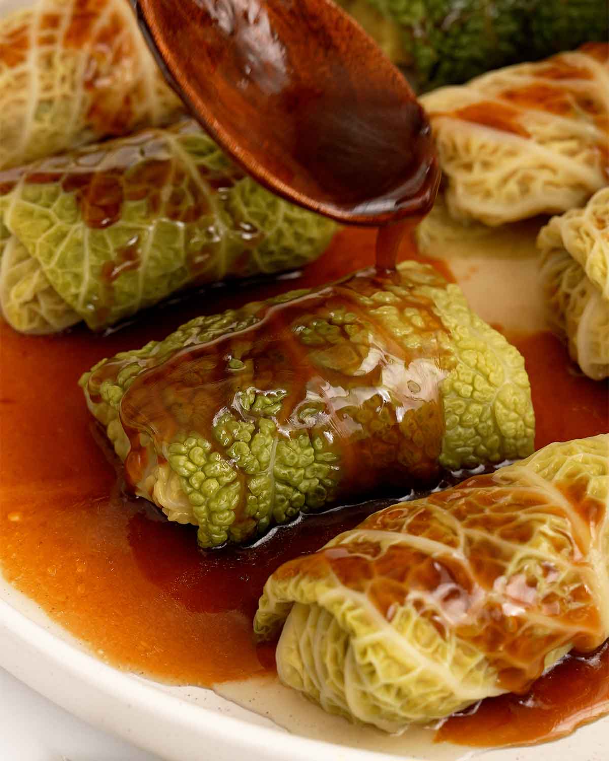 A white plate containing Chinese cabbage rolls and a wooden spoon pouring glossy sauce over the rolls.