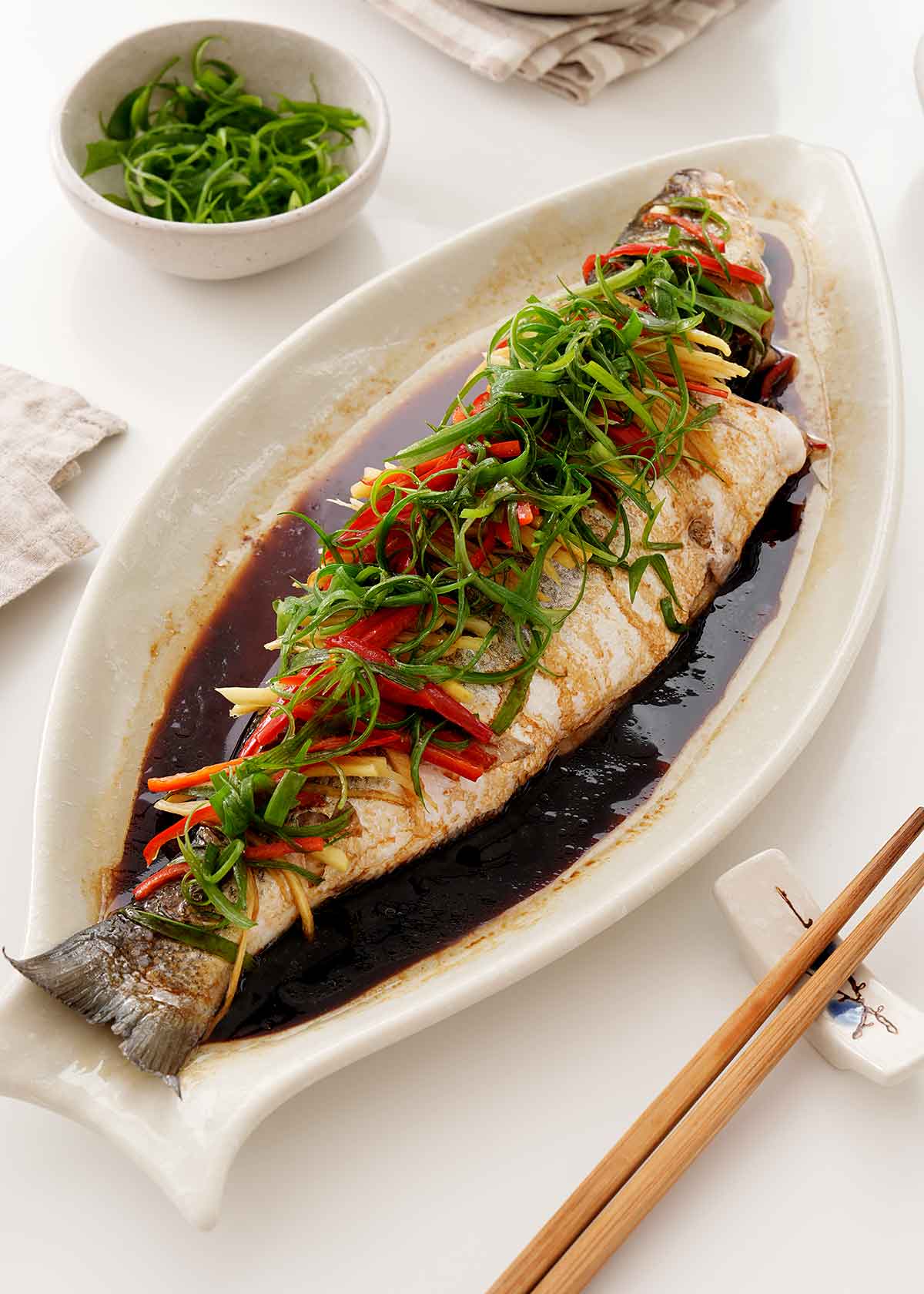 A large white oval plate containing steamed whole fish topped with green onions and chilli slices.