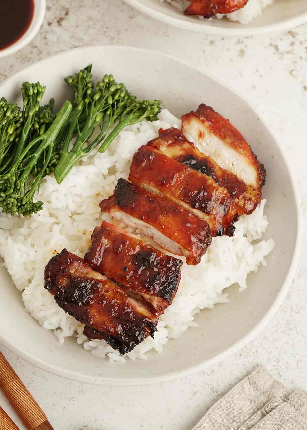 A white bowl containing rice and red char siu chicken slices with broccoli on the side.