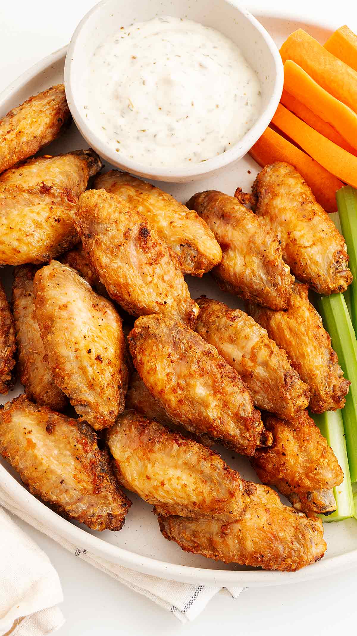 A white plate containing golden crispy skin fried chicken wings with celery, carrots and ranch sauce on the side.