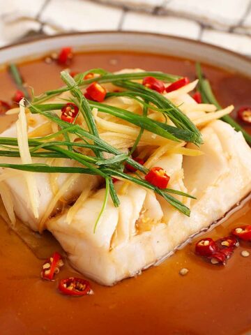 A white plate containing steamed white cod fish, garnish with green onions, red chillies, and ginger slices and brown colour seasoning sauce.