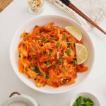 A white bowl containing ribbon texture carrot salad with spring onions and crushed peanuts and sesame seeds on top.