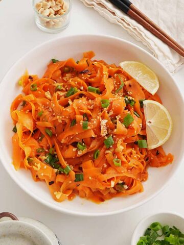 A white bowl containing ribbon texture carrot salad with spring onions and crushed peanuts and sesame seeds on top.