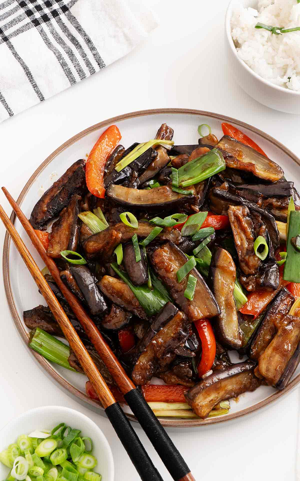 A white plate containing stir fried aubergine with red bell peppers slices and spring onions with a pair of chopsticks on the side.
