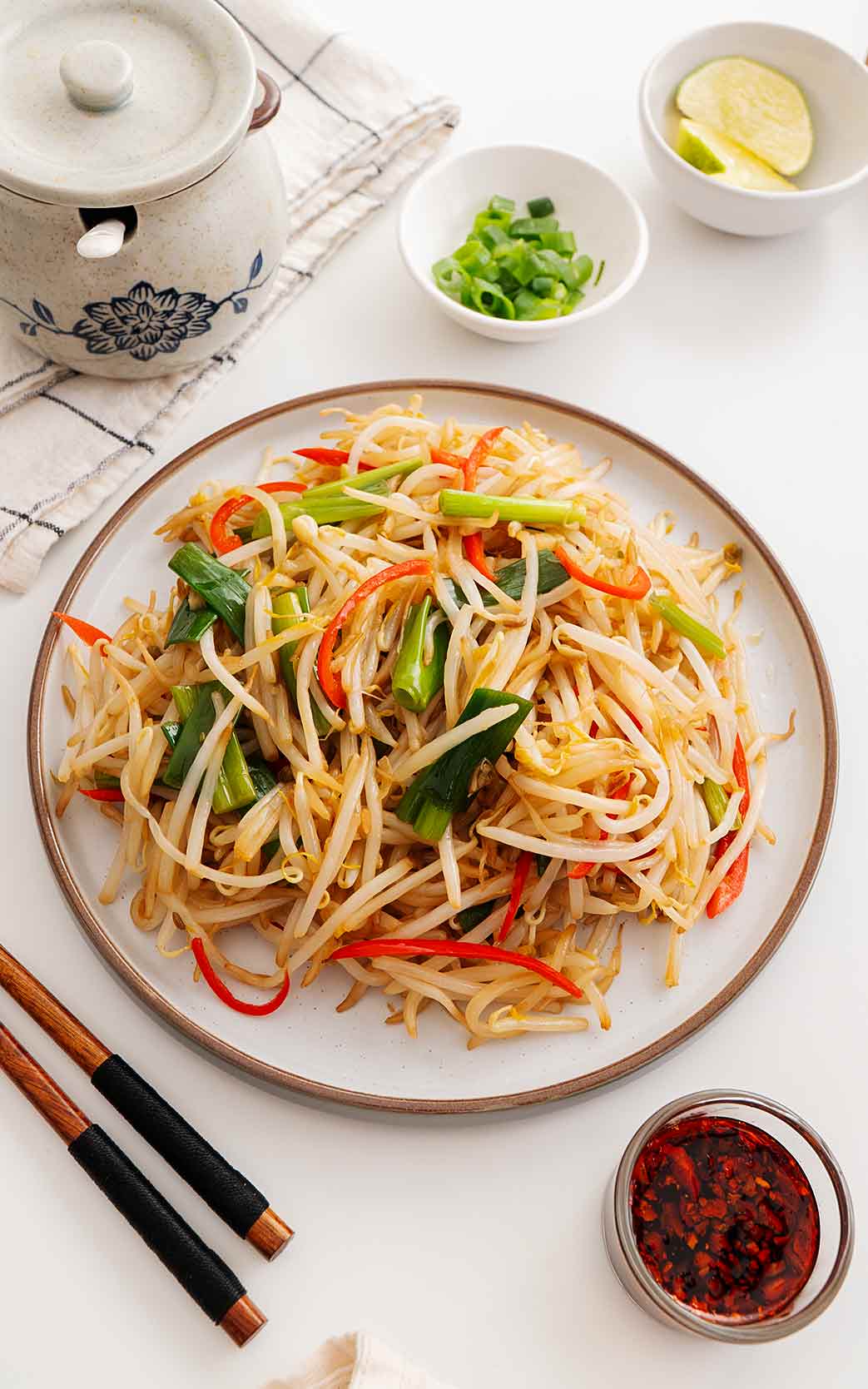 An image of a white plate containing fried beansprouts dish with chilli oil and chopsticks on the side.