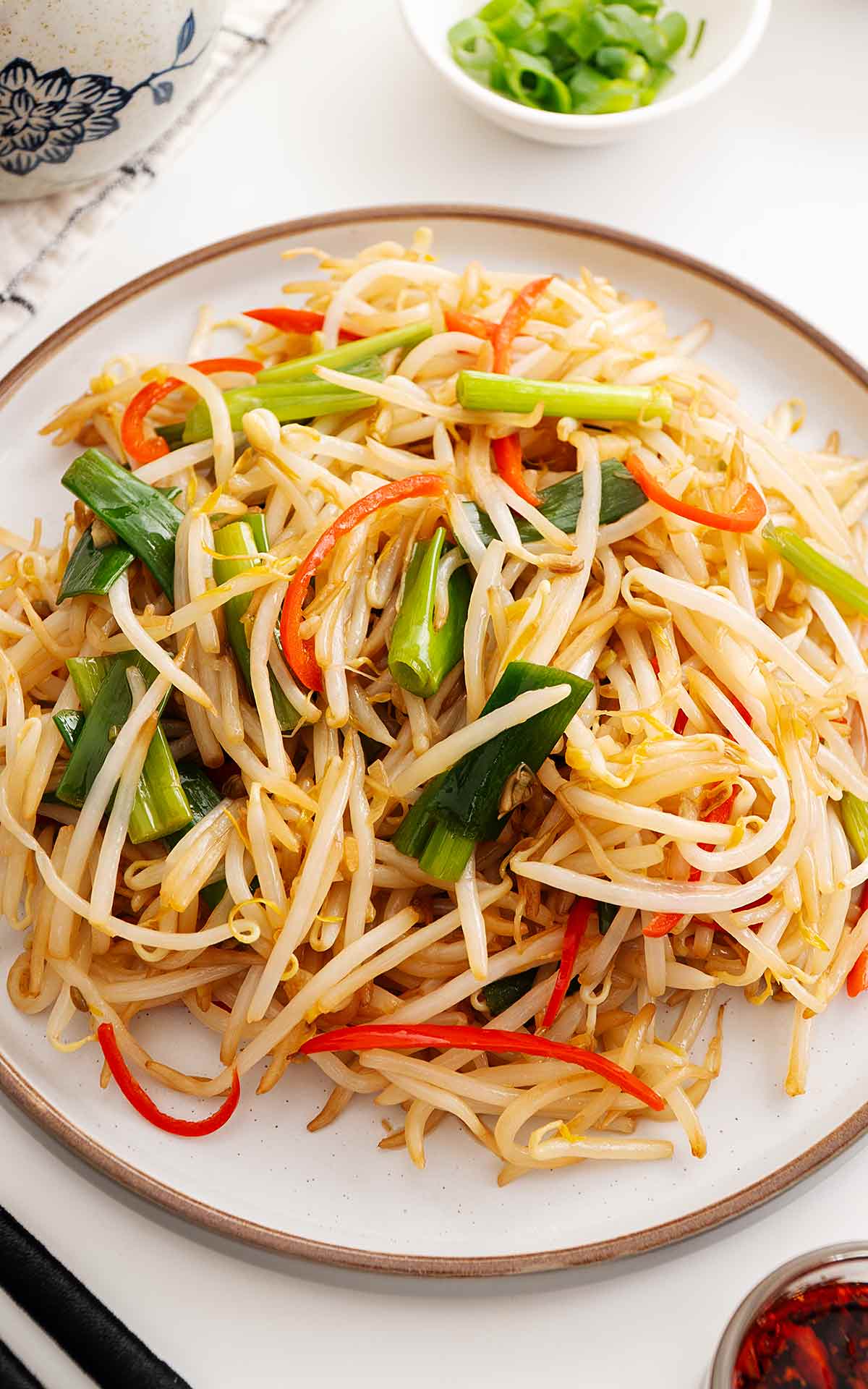 A white plate containing beansprout stir fry with green onions and red chillies.