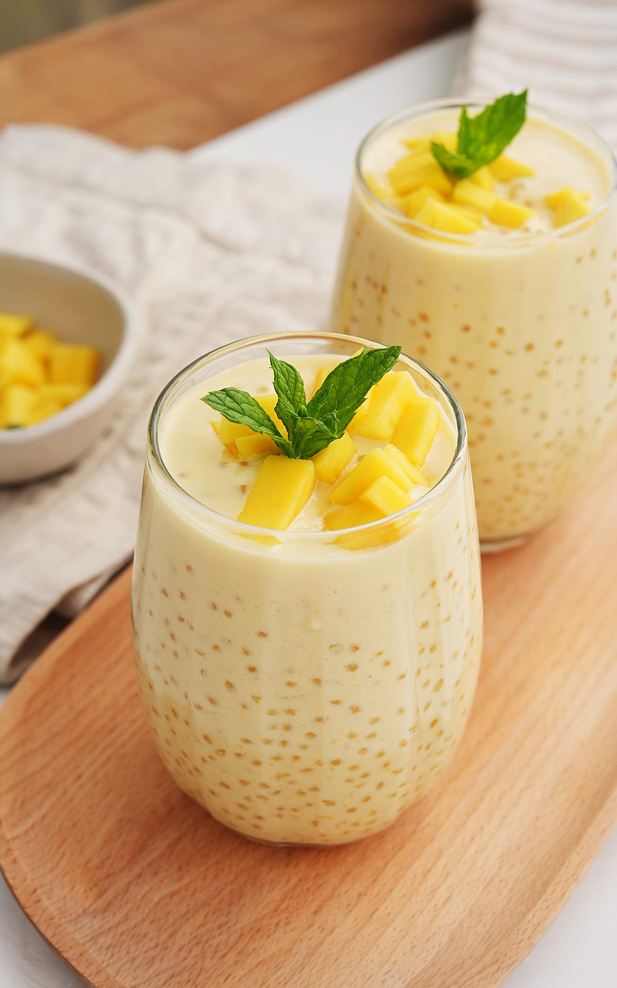 A glass cup containing a creamy sago drink with mango chunks and garnished with mint leaves.