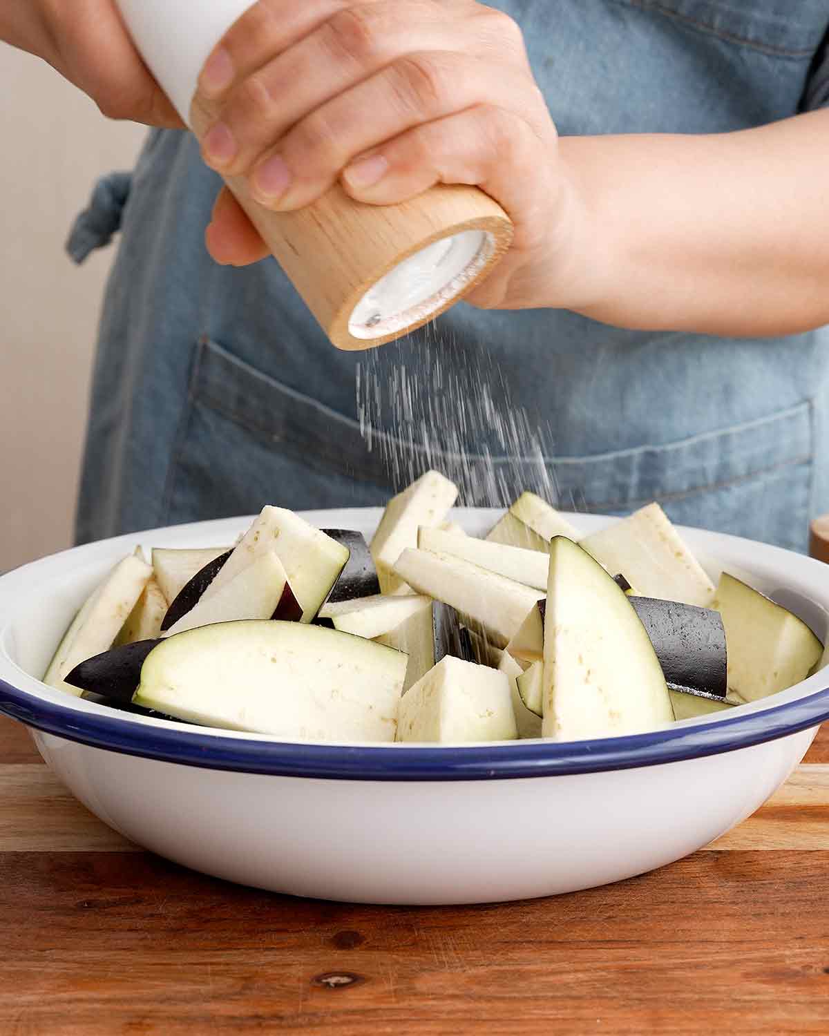 Large white bowl containing aubergine slices and sprinkling salt over it.