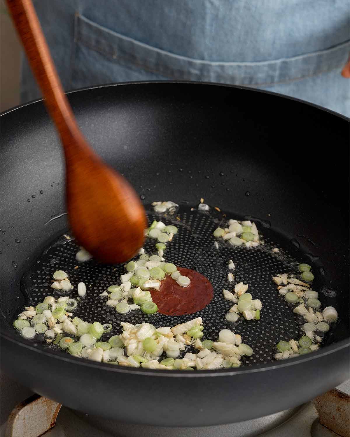 An image showing how to stir fry garlic and spring onions in a large wok.