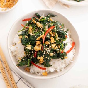 A white bowl containing white rice topped with spinach stir fry and garnished with fried garlic and sesame seeds.
