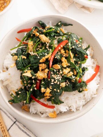 A white bowl containing white rice topped with spinach stir fry and garnished with fried garlic and sesame seeds.